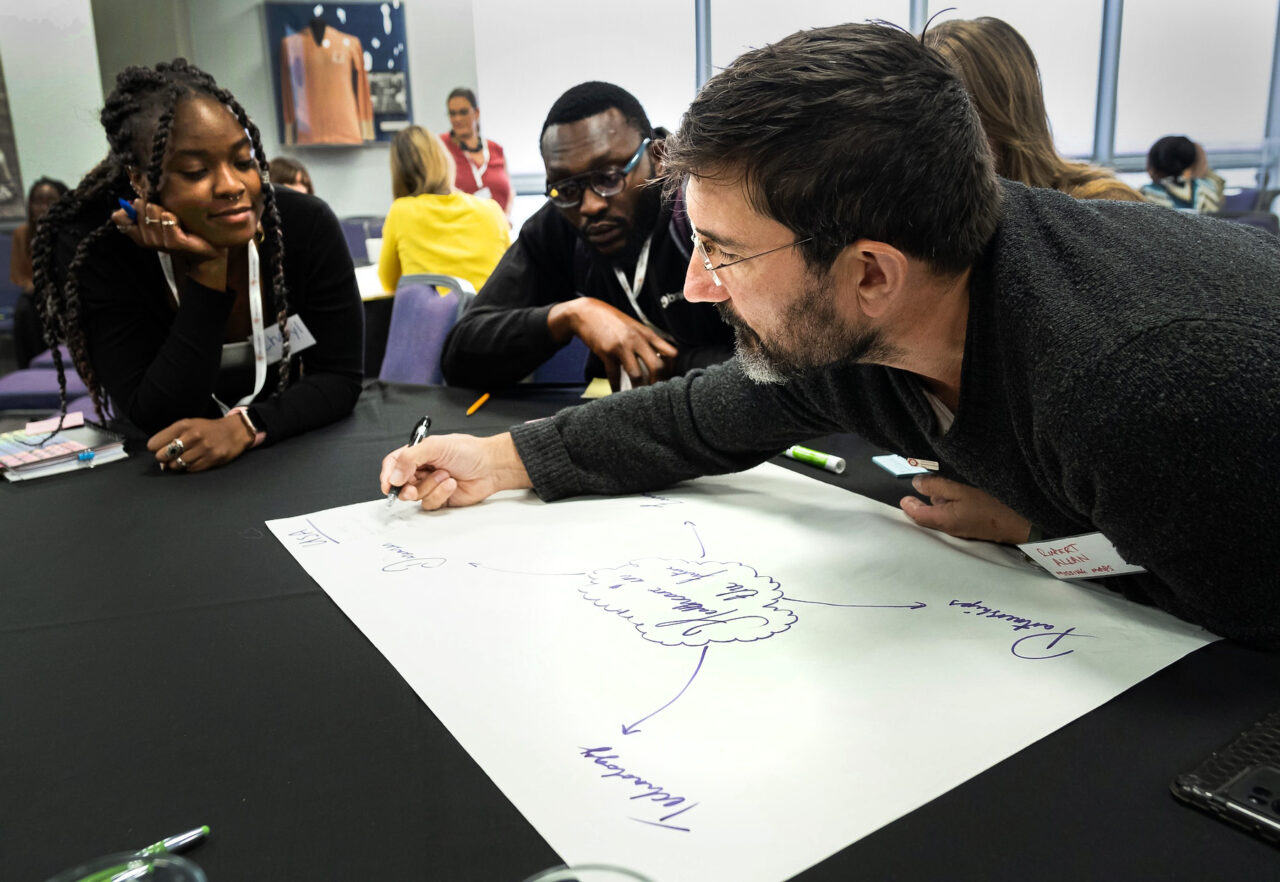 A group of people collaborate at a conference table, focused on a brainstorming session, with a man writing ideas on a large paper.
